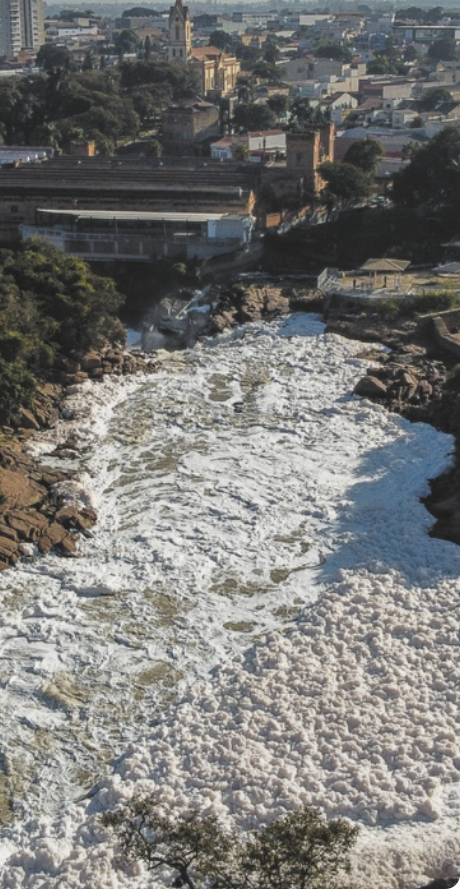 Fotografia de rio com grande quantidade de espuma. Ao fundo, uma cidade pequena.