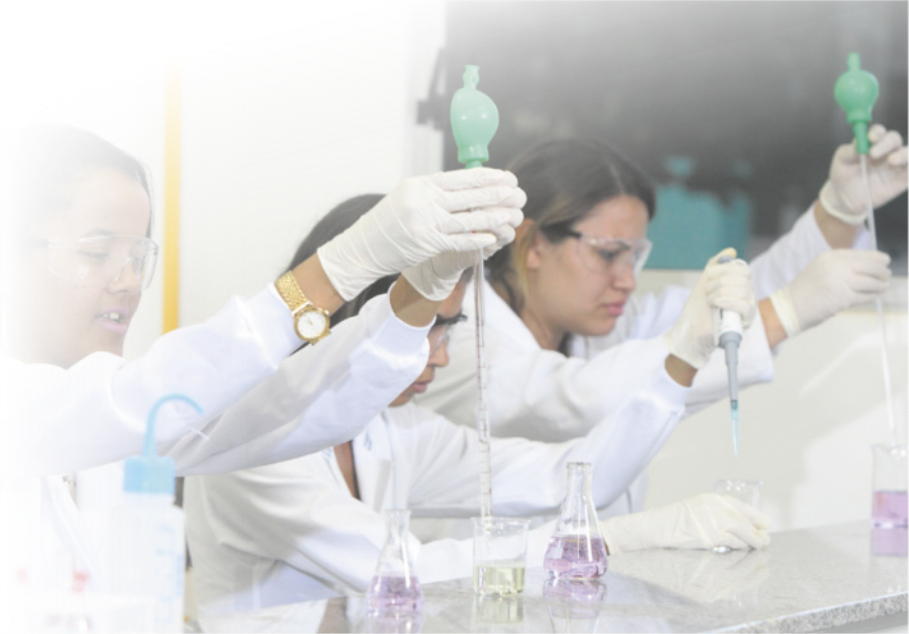 Imagem de pessoas sentadas em frente a uma bancada de laboratório. Elas vestem jalecos, óculos de proteção, luvas e manuseiam equipamentos de laboratório.