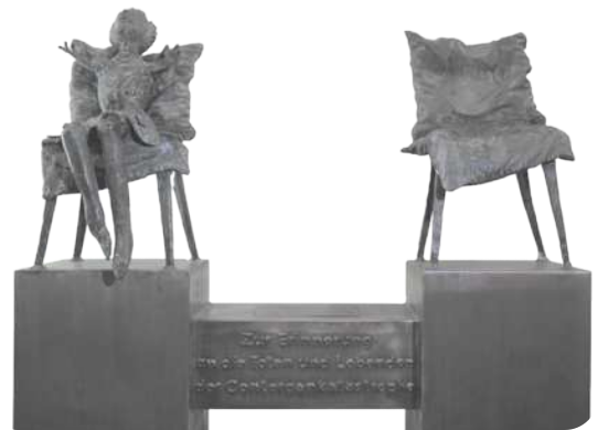 Fotografia de uma escultura com cadeiras. Em uma delas há uma pessoa com malformações nos membros. A outra está vazia.
