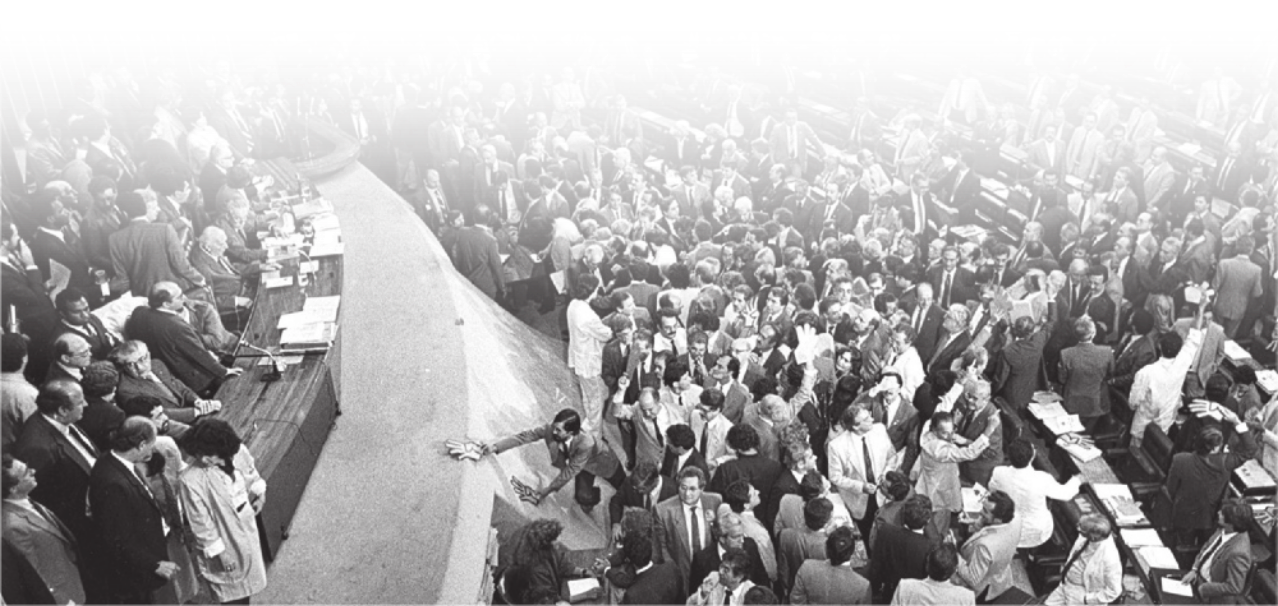 Fotografia aérea de pessoas no Congresso Nacional. Em uma região elevada, há uma bancada onde algumas pessoas estão posicionadas. O local está repleto de pessoas usando roupas sociais, como ternos e gravatas.