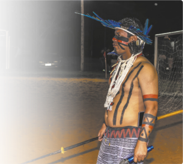 Fotografia de um homem indígena com um cocar de penas, um colar de cordões, bermuda e pinturas corporais nas cores preto e vermelho. Ele segura uma lanterna em uma das mãos e um bastão adornado na outra. Ao fundo, um campo de futebol de areia.