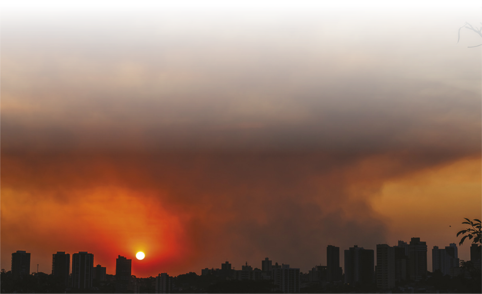 Fotografia de área urbana com muita fumaça no céu, com o sol parcialmente coberto por ela.