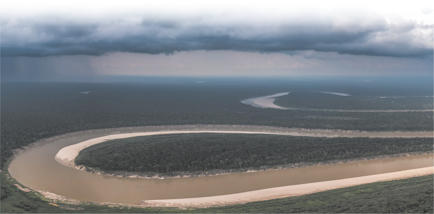 Fotografia aérea de um rio em uma região de floresta. O rio forma curvas e o céu está encoberto por nuvens. É possível observar chuva em algumas regiões.