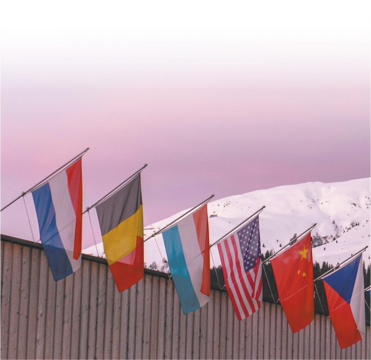 Fotografia de bandeiras de países alinhadas no topo de uma construção em uma região de montanha e neve.