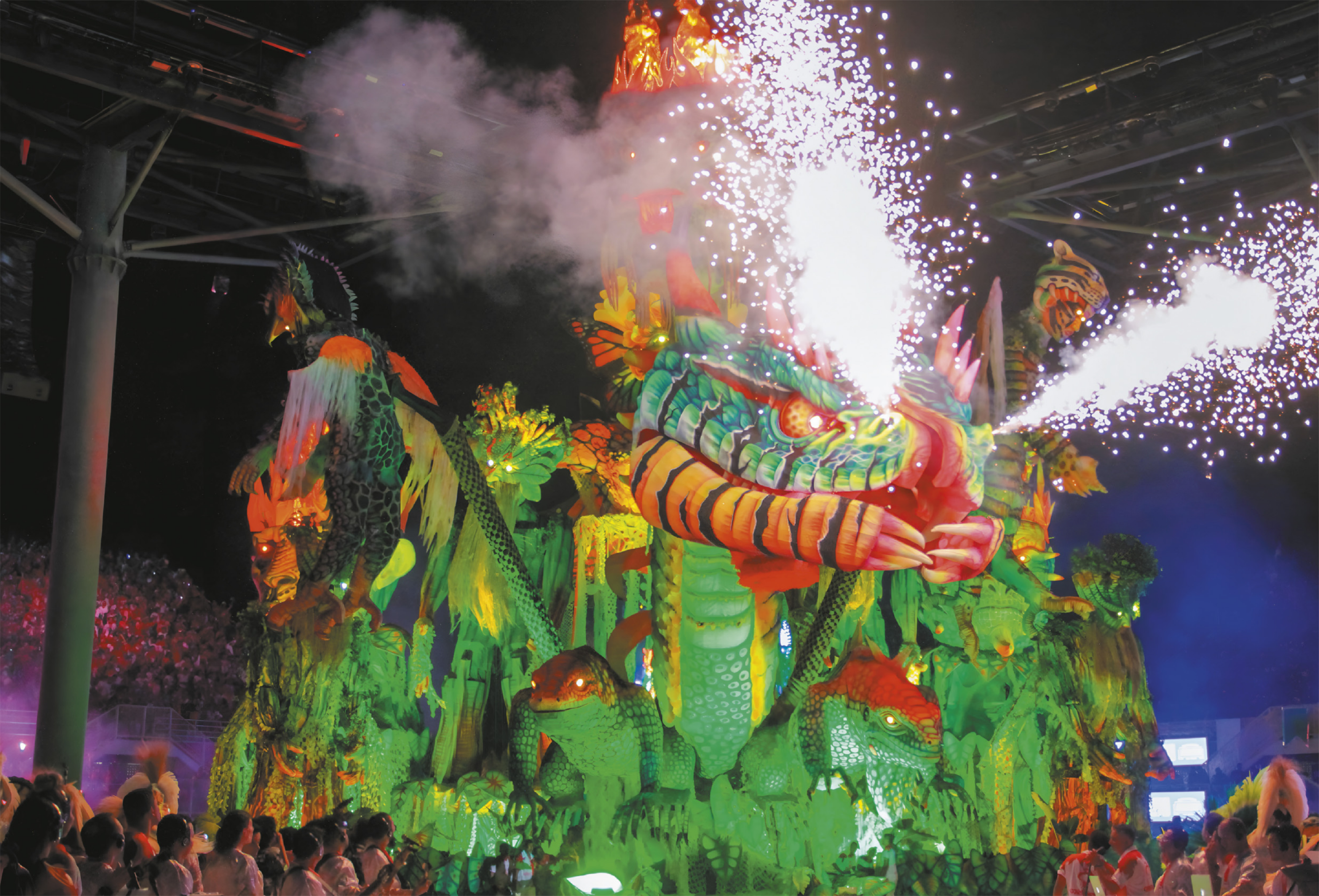  Fotografia de um carro alegórico rodeado por pessoas. Sua estrutura principal tem o formato de um dragão, com fumaça e faíscas saindo de suas narinas. Ao redor do dragão, há diversas outras estruturas com formas de animais reptilianos.