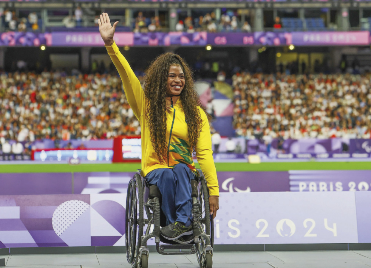 Fotografia de Raissa Machado: uma mulher com cabelos longos e cacheados, sorrindo com um braço erguido para o alto, sentada em uma cadeira de rodas. Ao fundo há uma arquibancada com uma plateia em torno de uma pista de atletismo.