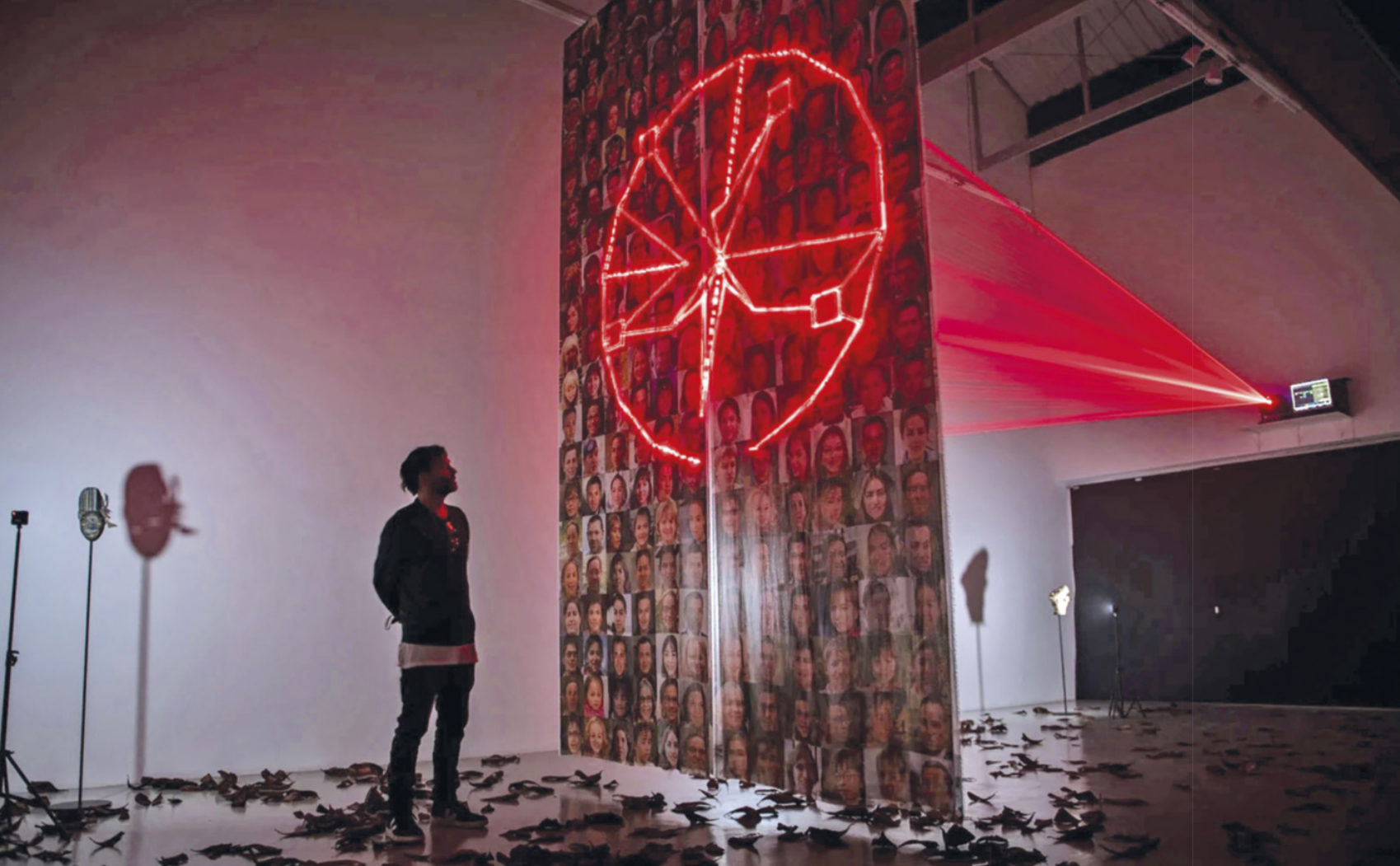 Fotografia de Fernando Velázquez: um homem de barba e cabelos curtos em pé diante de uma instalação artística com a forma de uma grande faixa de tecido pendendo do teto de uma sala expositiva, com muitos rostos de pessoas impressos no tecido e um desenho geométrico de formato circular, com linhas, quadrados e triângulos internos ao círculo, projetado sobre os rostos na faixa por um feixe de lasers, que é propagado a partir de um aparelho instalado no alto de uma parede da sala. O chão da sala está repleto de folhas secas espalhadas pelo espaço.