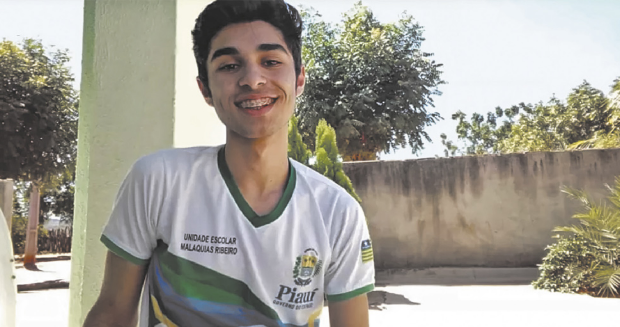 Fotografia de Samuel Castro: um garoto sorridente com aparelho fixo nos dentes e cabelos curtos, usando um uniforme escolar, com um muro e árvores ao fundo.