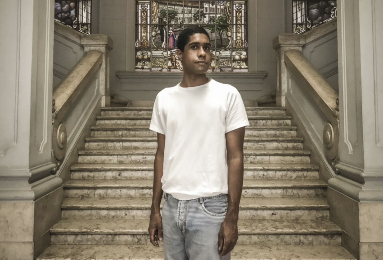 Fotografia de Juan Comamala: um homem em pé em frente a uma escadaria de mármore em um edifício de arquitetura neoclássica. Ele tem cabelos curtos e está vestido com uma camiseta e calça jeans.