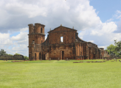 Fotografia de um edifício em ruínas com arquitetura do período colonial brasileiro. Ele está situado em uma paisagem com um gramado e árvores ao fundo.