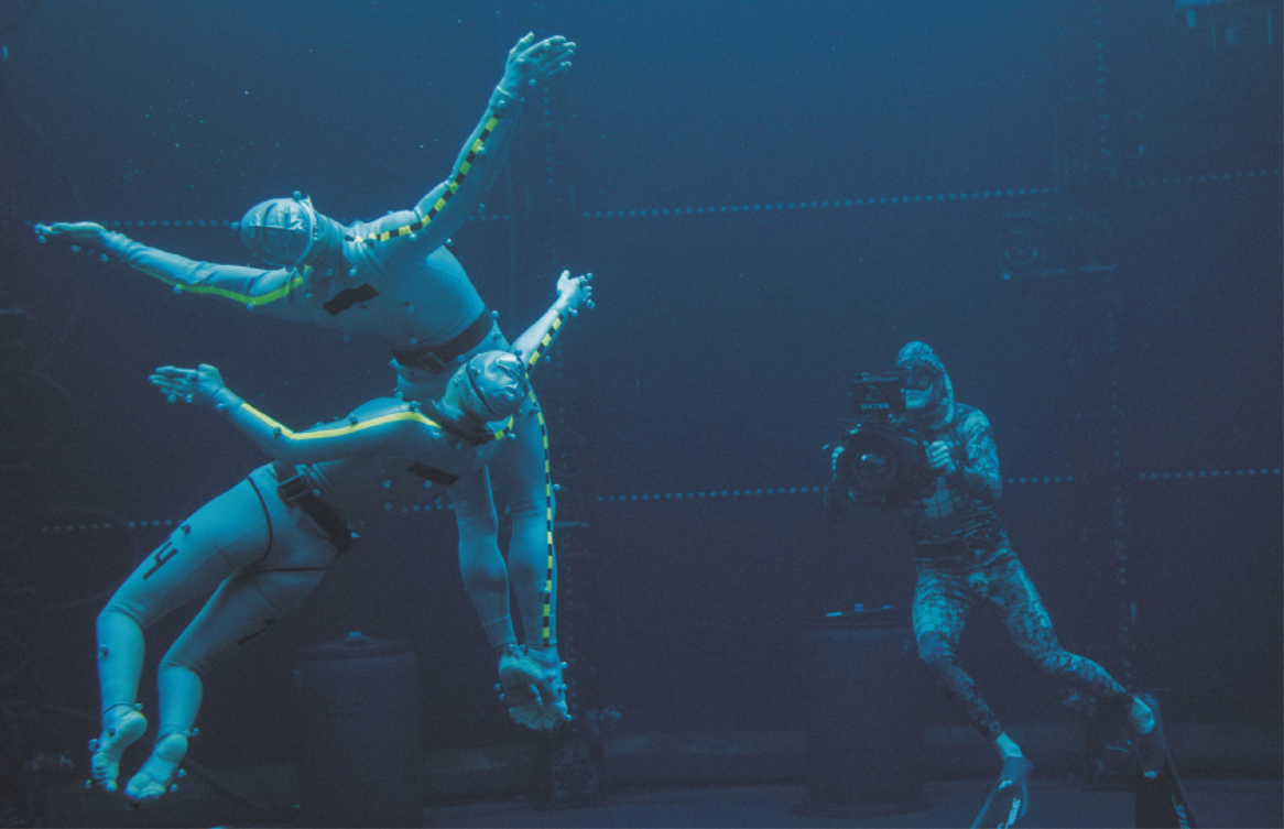 Fotografia de atores nadando e executando uma coreografia de dança, submersos em um tanque, vestidos com roupas que cobrem todo o corpo, com pequenas esferas instaladas em diversos pontos da superfície do corpo. Ao fundo há um homem com uma câmera nadando no fundo do tanque, usando equipamentos de mergulho e filmando-os com uma câmera.