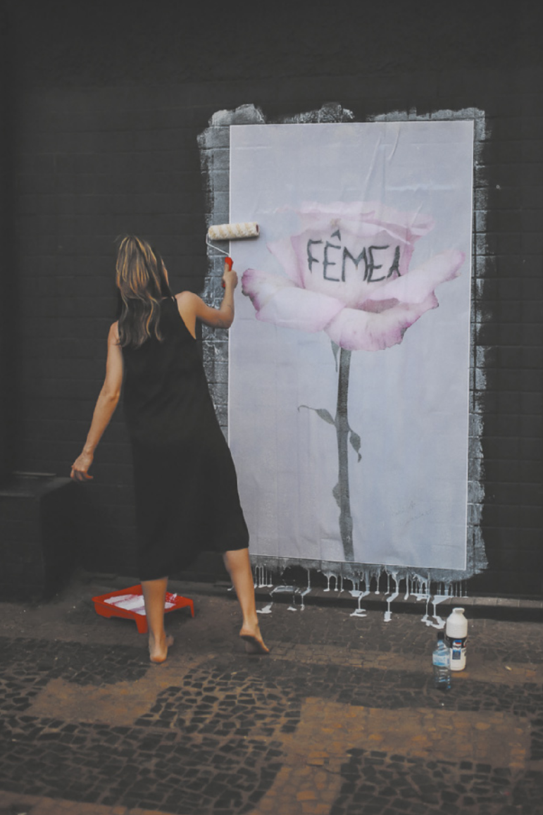 Fotografia de uma mulher de vestido colando um cartaz em uma parede, usando um rolo com cola branca. O cartaz mostra a imagem de uma rosa com uma pétala em que está escrito 'fêmea'.