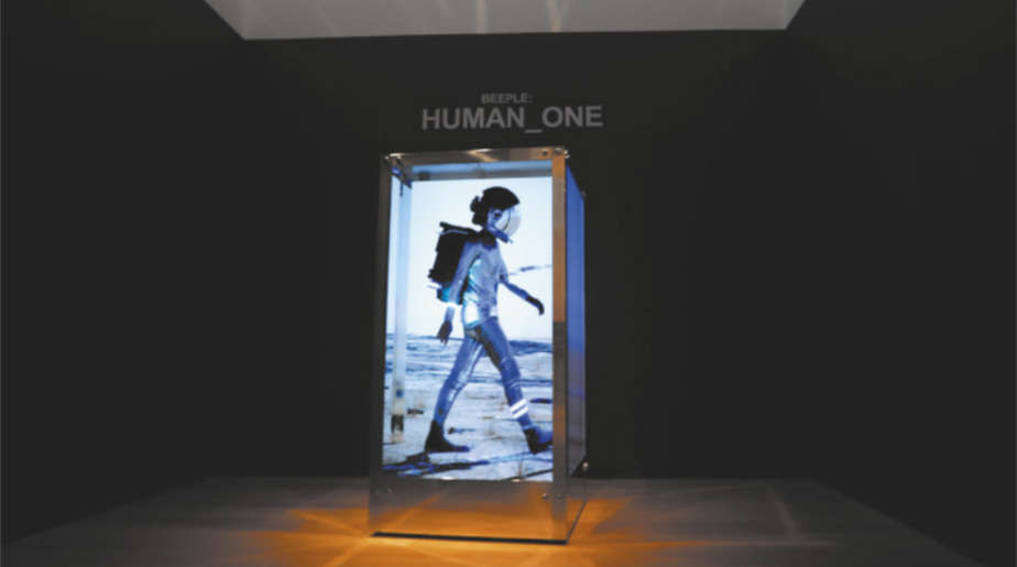 Fotografia de uma escultura no centro de um espaço expositivo escuro, com a forma de uma grande caixa retangular metálica, com telas luminosas nas faces da caixa mostrando a imagem de uma pessoa caminhando com roupas brilhantes e um capacete de astronauta, carregando uma mochila, em uma paisagem árida.