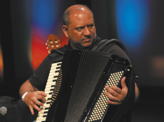 Fotografia de Marcelo Cigano. Ele é um homem calvo, tocando um acordeom.