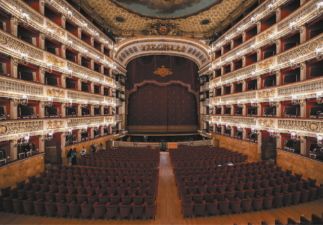 Fotografia do interior de um teatro com uma plateia central em frente ao palco e cinco andares de camarotes de plateias laterais. O palco está coberto por cortinas e o teto do teatro é decorado com pinturas e ornamentos.
