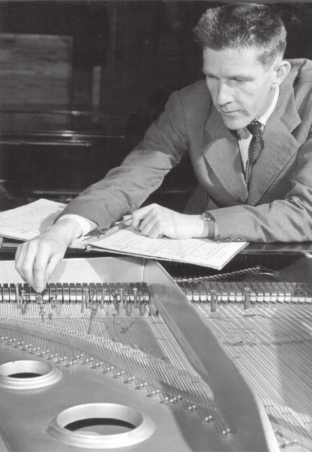 Fotografia em preto e branco de John Cage. Ele é um homem com cabelos curtos, vestido com terno e gravata, manuseando as cordas de um piano de cauda, segurando uma pasta com folhas de papel com partituras.