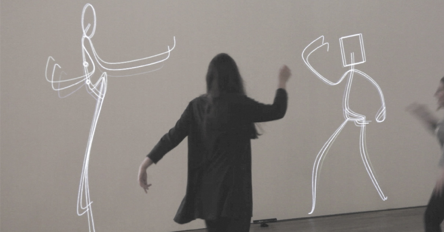 Fotografia de uma mulher de costas dançando diante de figuras humanas esquemáticas, desenhadas com linhas luminosas projetadas sobre uma parede. As figuras estão realizando movimentos de dança e têm a escala de um ser humano.