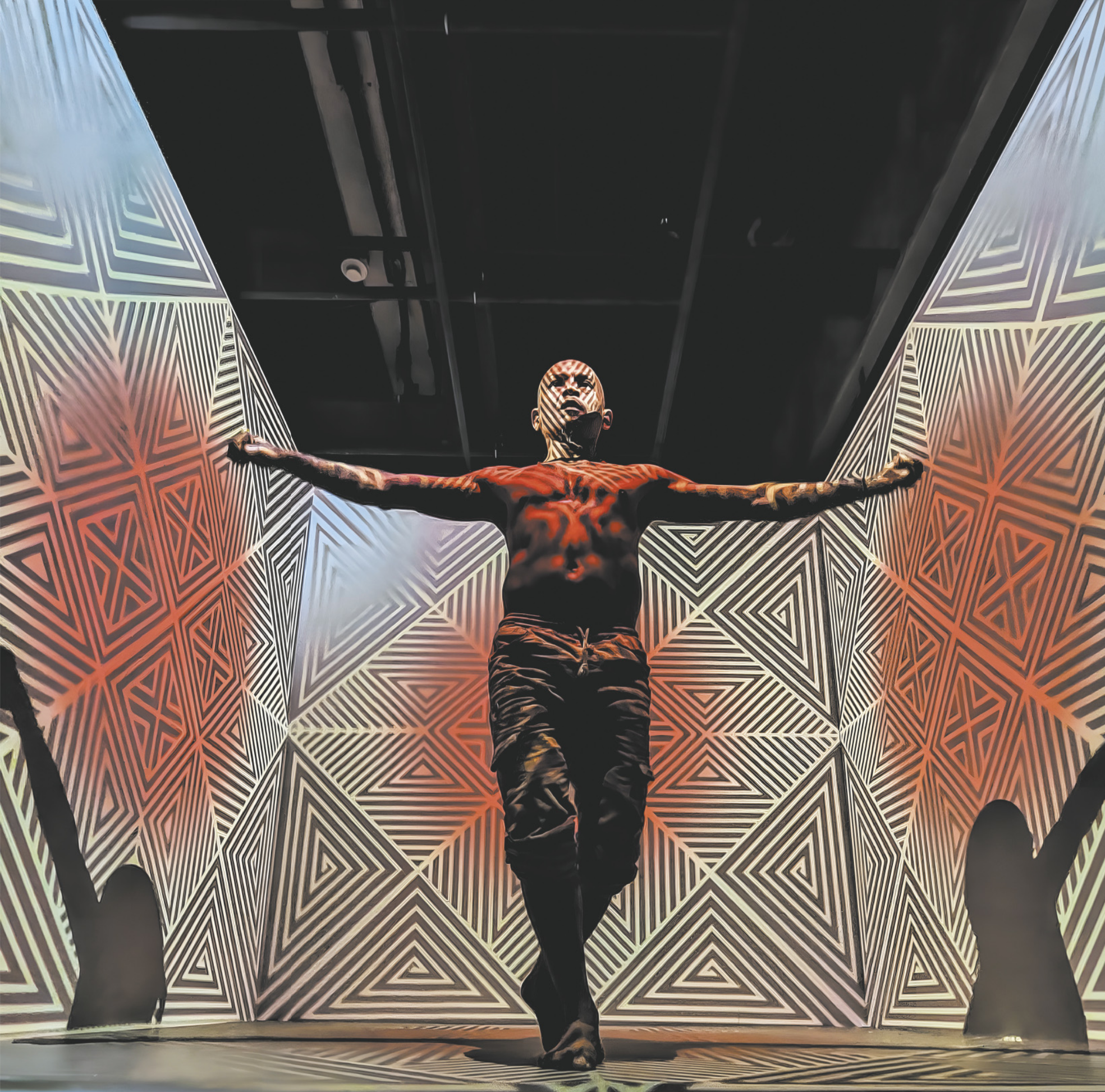 Fotografia de um homem realizando uma performance em uma instalação com grafismos geométricos de formas triangulares e de losangos projetados nas paredes e no chão de um ambiente expositivo. Ele está em pé, sem camiseta e de braços abertos, e os grafismos também se projetam sobre o corpo dele.
