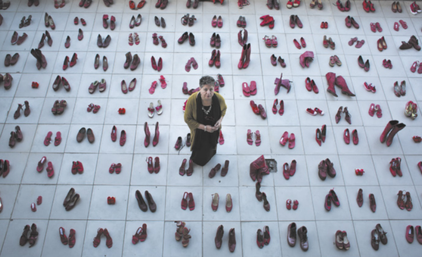 Fotografia de Elina Chauvet em pé sobre uma instalação artística em que há centenas de pares de sapatos vermelhos organizados, com cada par em um quadrado no chão.