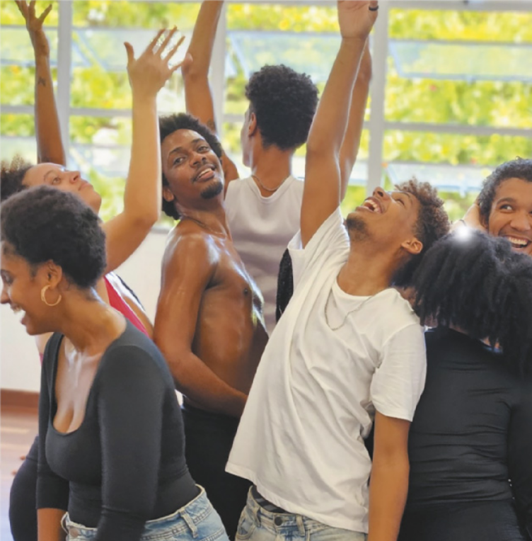 Fotografia de um grupo de atores jovens, sorrindo com os corpos próximos e os braços erguidos para o alto, em uma sala.