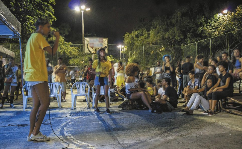 Fotografia de duas mulheres em pé segurando microfones na mão, diante de uma plateia de pessoas sentadas no chão e em cadeiras e bancos, em uma quadra esportiva, à noite.
