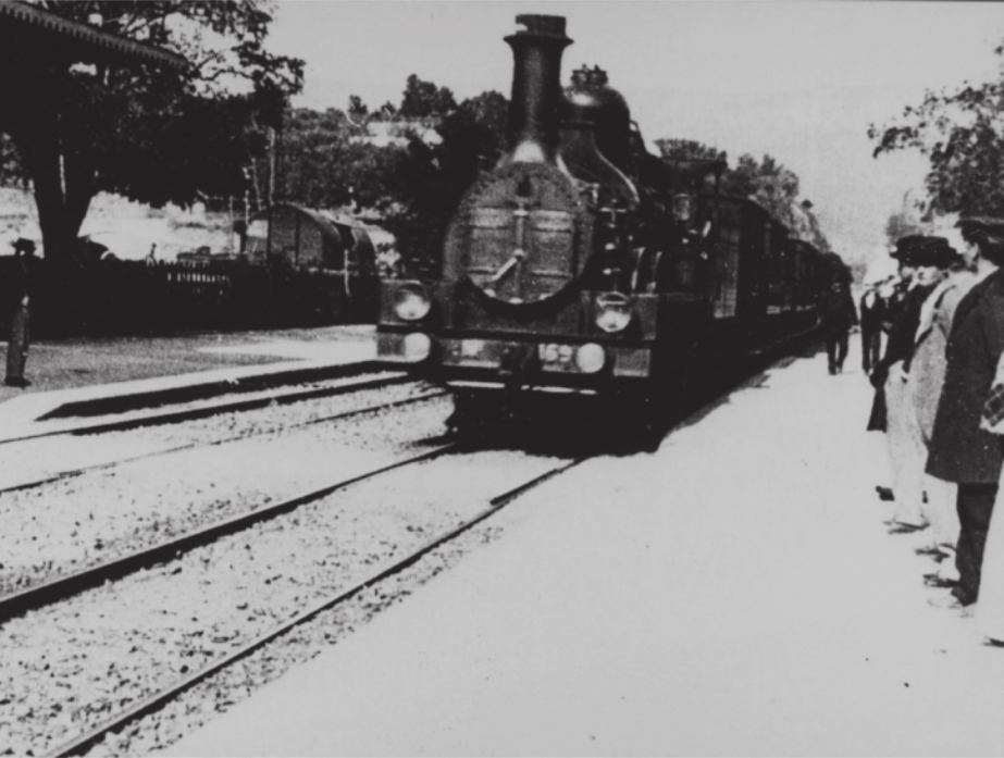 Fotografia em preto e branco de um trem em movimento sobre um dos trilhos de uma estrada de ferro, com um grupo de homens ao lado do trilho, observando.