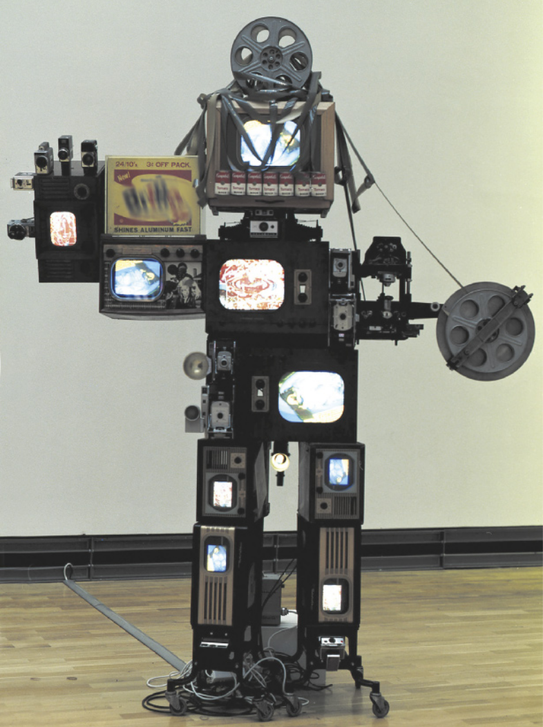 Fotografia de escultura representando um robô com o corpo feito de diversos tipos de televisores, com as telas mostrando imagens, e equipamentos eletrônicos, além de outros objetos descartados. Há partes do corpo do robô com câmeras, projetores, caixas de papelão, latas de sopa, cabos e rolos de filme.