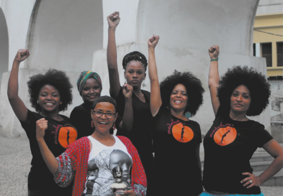 Fotografia de um grupo de seis mulheres com os punhos fechados erguidos para o alto. Cinco delas estão uniformizadas; três delas têm cabelos cacheados longos; uma delas têm o cabelo curto e está de óculos; outra está usando um lenço nos cabelos; e outra está com os cabelos presos.