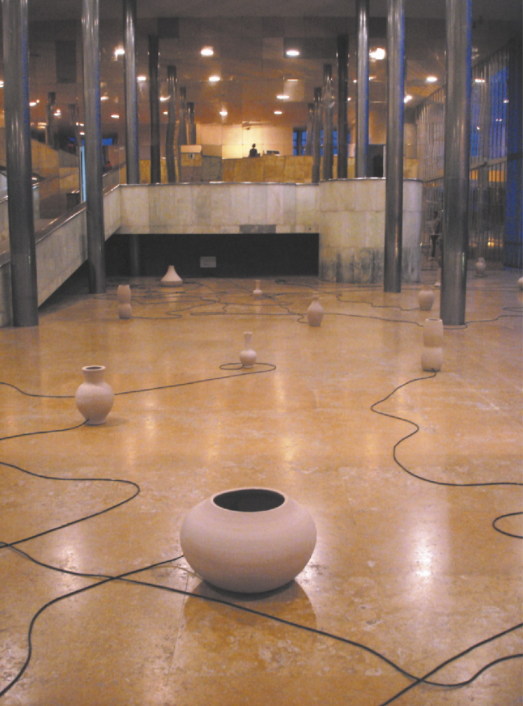Fotografia de instalação artística com vasos e jarras de cerâmica espalhados pelo piso do interior de um edifício de arquitetura modernista. Os vasos têm pequenos orifícios por onde passam cabos elétricos, dispostos pelo espaço entre as peças de cerâmica como linhas ondulantes.