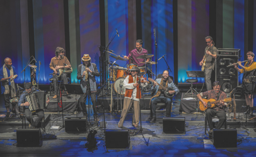Fotografia de um grupo de músicos em um palco, tocando diversos instrumentos musicais como rabeca, viola caipira, banjo, bateria, flauta transversal, baixo elétrico, clarinete, pandeiro e acordeom.