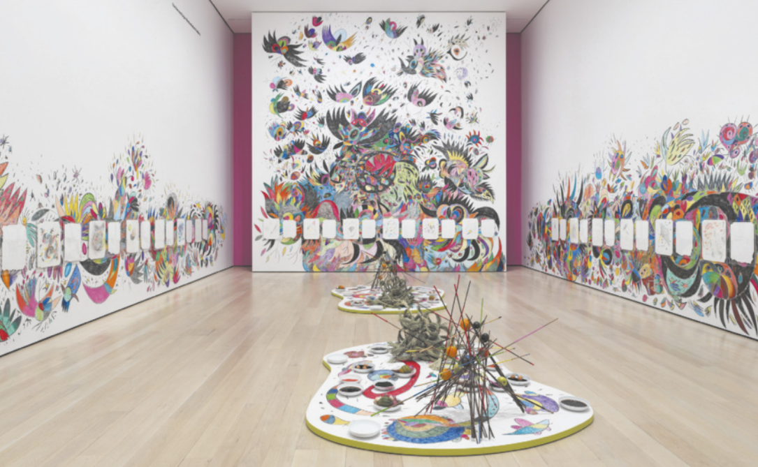 Fotografia de instalação artística em um ambiente expositivo com desenhos coloridos abstratos de formas curvas e arredondadas e figuras semelhantes a pássaros nas paredes, além de desenhos em folhas brancas expostos nas paredes e duas esculturas feitas com varetas de madeira aglomeradas e justapostas, expostas sobre painéis no chão, também contendo desenhos e pinturas, ao lado de pratos com alimentos.