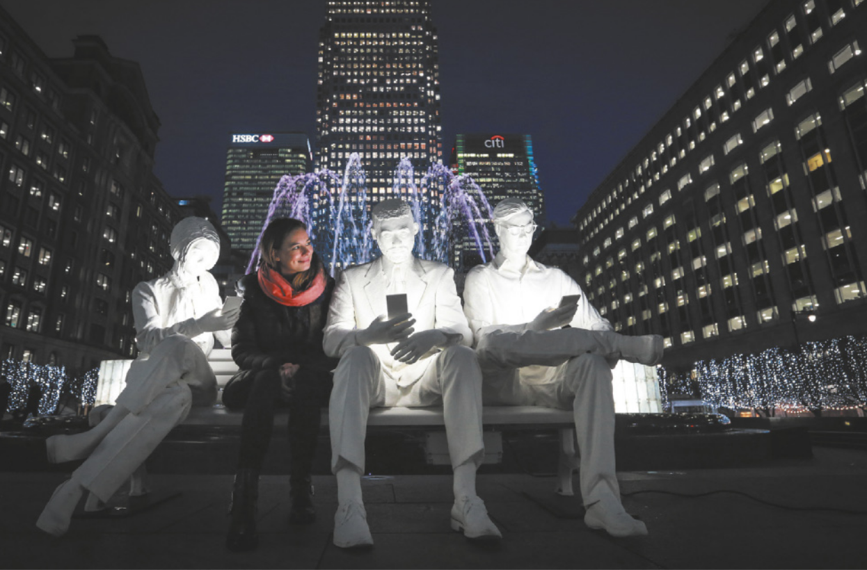 Fotografia de uma mulher sentada em um banco de praça, ao lado de esculturas representando pessoas sentadas olhando para o smartphone na mão. Das telas dos smartphones emana uma luz que ilumina as esculturas e, ao fundo, há um chafariz com águas iluminadas por luzes coloridas e edifícios corporativos com as janelas iluminadas, à noite.