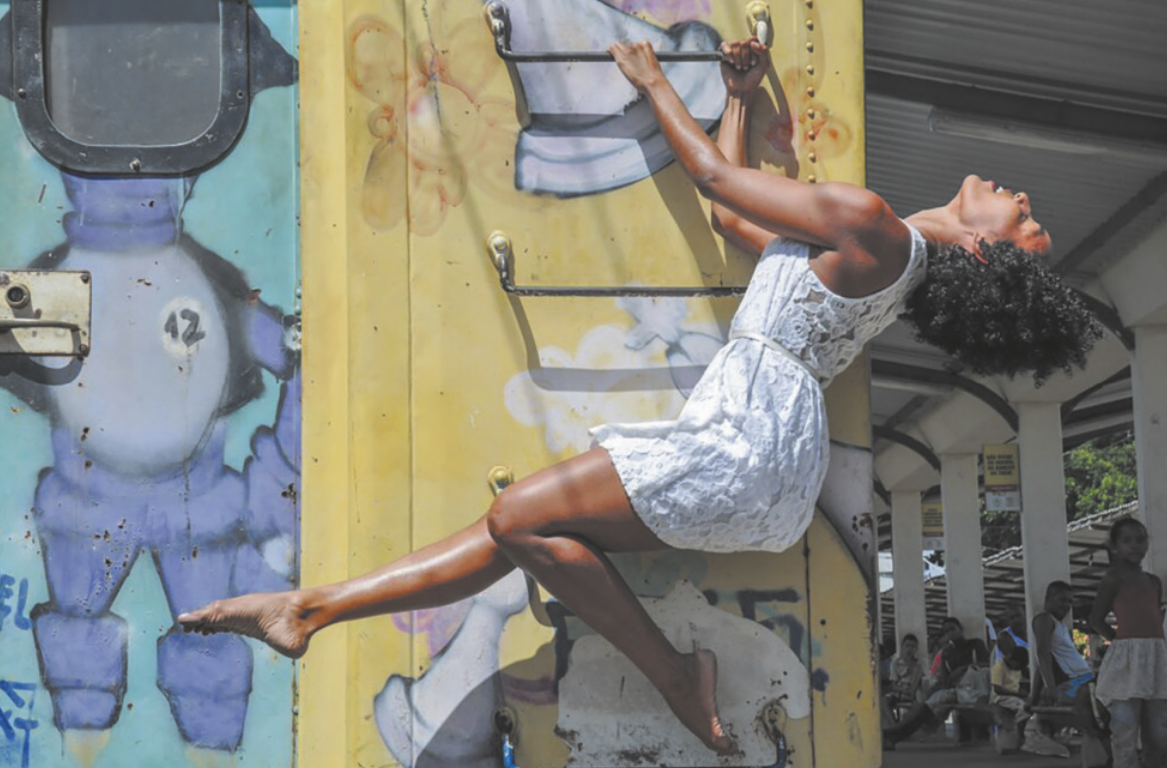 Fotografia de Deise Gabrielle. Ela é uma bailarina com cabelos longos e cacheados, usando um vestido. Ela está pendurada em uma escada vertical em uma parede, sustentando o corpo com as mãos, inclinando a cabeça para trás e estendendo uma das pernas para a frente, com a outra perna flexionada.