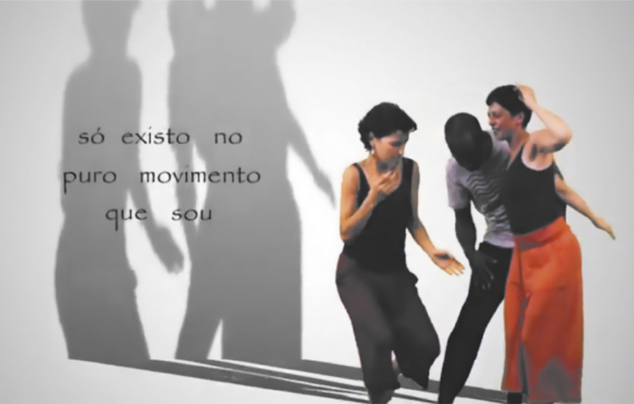 Fotografia de duas mulheres e um homem dançando, com as pernas flexionadas e os troncos inclinados, movimentando os braços. Eles estão sobre um pano de fundo branco, e a sombra deles se projeta sobre esse fundo, onde está escrita a mensagem 'só existo no puro movimento que sou'.