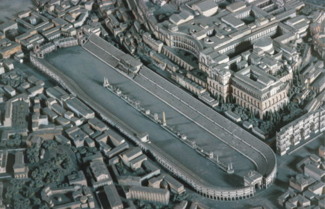 Fotografia de uma maquete representando uma cidade com muitos edifícios baixos e casas. No centro da cidade, há uma grande construção de formato alongado e arredondado nas extremidades, com uma arquibancada que percorre toda sua lateral. Na arena central da construção, há um canteiro central separando os dois lados da pista.