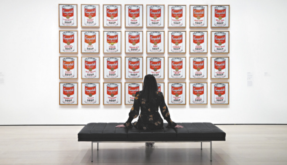 Fotografia de uma mulher sentada em um banco de um espaço expositivo diante de uma obra de arte exposta na parede. A obra consiste em 24 imagens emolduradas de uma lata de sopa pronta da marca Campbell's.