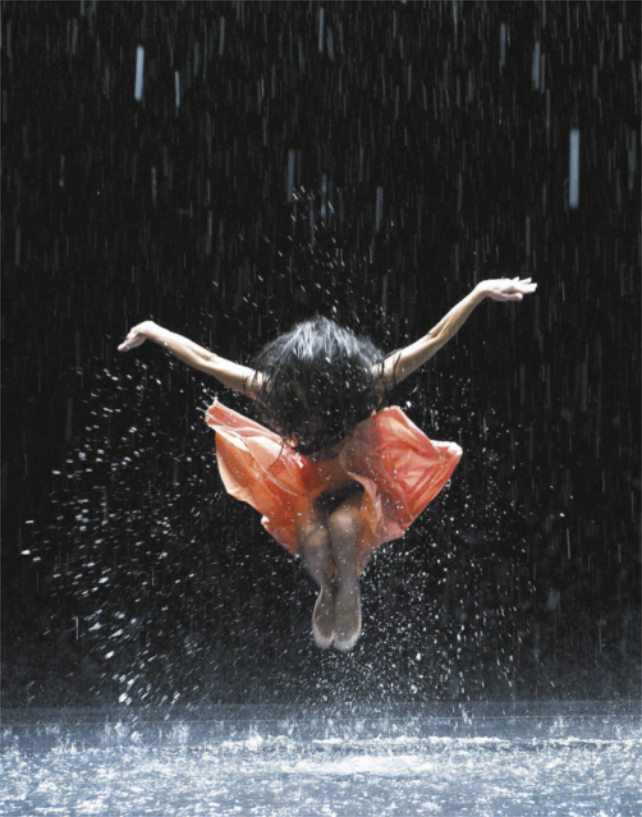 Fotografia de uma bailarina de saia, saltando no ar com os braços estendidos para os lados, à altura da cabeça, e as pernas unidas com os joelhos flexionados. Está chovendo sobre ela, e o chão está coberto de água, de modo que o salto da bailarina provoca respingos.