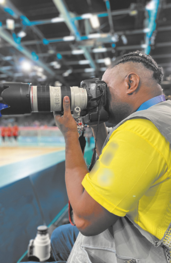Fotografia de um homem com cabelos presos em tranças curtas fotografando um evento esportivo em um ginásio, usando uma câmera fotográfica com a lente alongada.