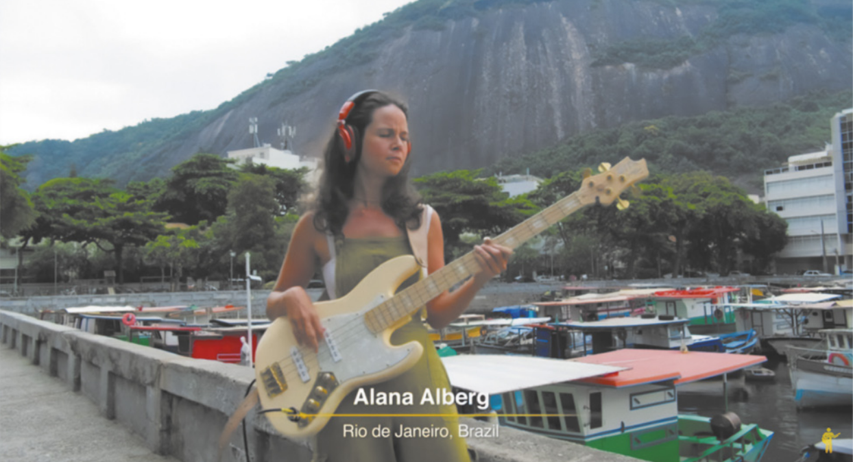 Fotografia de Alana Alberg. Ela é uma mulher de cabelos longos, usando fones de ouvido, tocando baixo elétrico. Ela está em pé em uma paisagem portuária, com barcos de pesca atracados a um cais atrás dela, árvores e um morro ao fundo.