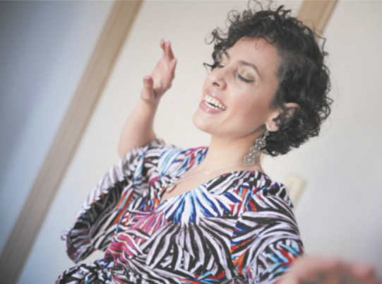 Fotografia de Aline Miklos. Ela é uma mulher de cabelos curtos e cacheados, usando brincos e uma camisa colorida, cantando com os braços erguidos ao lado do corpo.