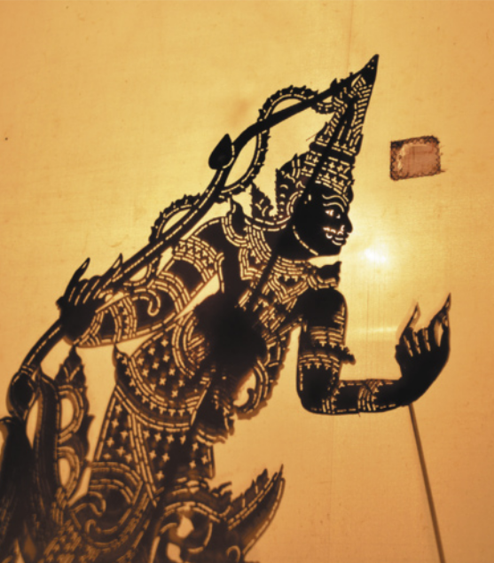 Fotografia de um fantoche representando uma figura mitológica vestida com uma espécie de armadura com muitos detalhes e ornamentos. O fantoche é feito com papéis articulados ricos em detalhes e ornamentos vazados, por onde a luz ao fundo passa, fazendo com que o fantoche apareça como uma silhueta escura sobre um fundo luminoso.