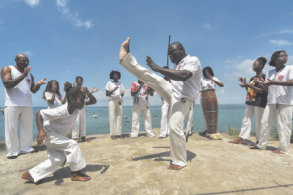 Fotografia de uma roda de capoeira à beira do mar, com pessoas em pé, batendo palmas e tocando instrumentos. Ao centro, uma menina esquiva de um homem, que chuta o ar com o pé à altura da cabeça.