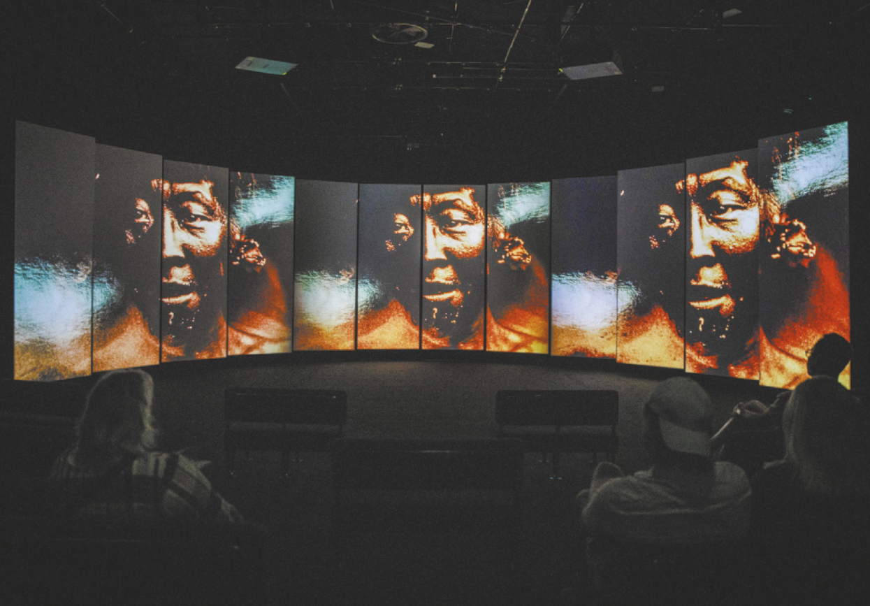 Fotografia de pessoas em um espaço expositivo escuro sentadas em cadeiras, assistindo a um vídeo exibido em um painel composto por 12 telas de formato alongado, na vertical, justapostas em uma disposição semicircular. As telas mostram três vezes uma mesma fotografia de um homem indígena, sobrepostas à fotografia de reflexos na água.