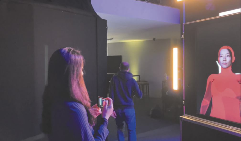 Fotografia de uma mulher em um espaço expositivo, apontando a câmera do smartphone em direção à tela de uma obra de arte interativa, que faz com que o rosto da mulher apareça refletido na tela, porém vestida com uma espécie de macacão colorido, com um capuz na cabeça.