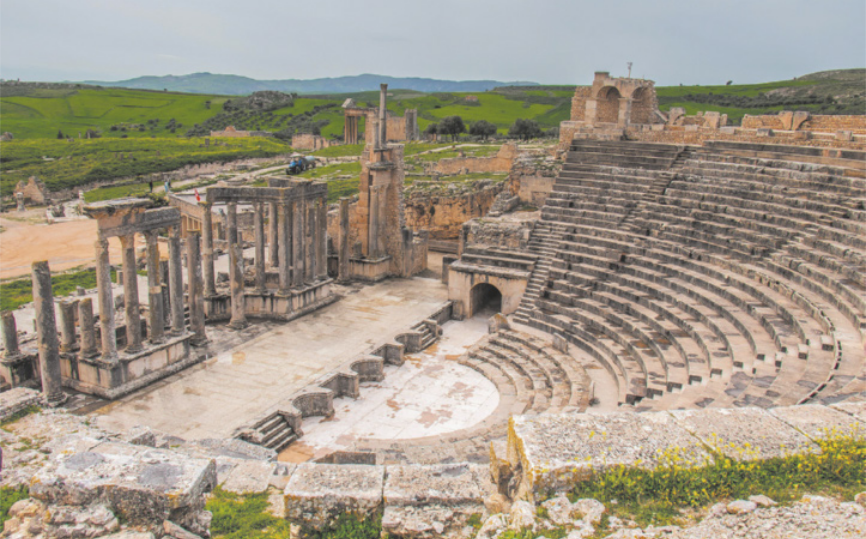 Fotografia de ruínas de um teatro com arquibancadas de formato semicircular, diante de um palco retangular. Na parte de trás do palco, há uma construção deteriorada, formada por colunas. Ao fundo há uma paisagem de morros e colinas.