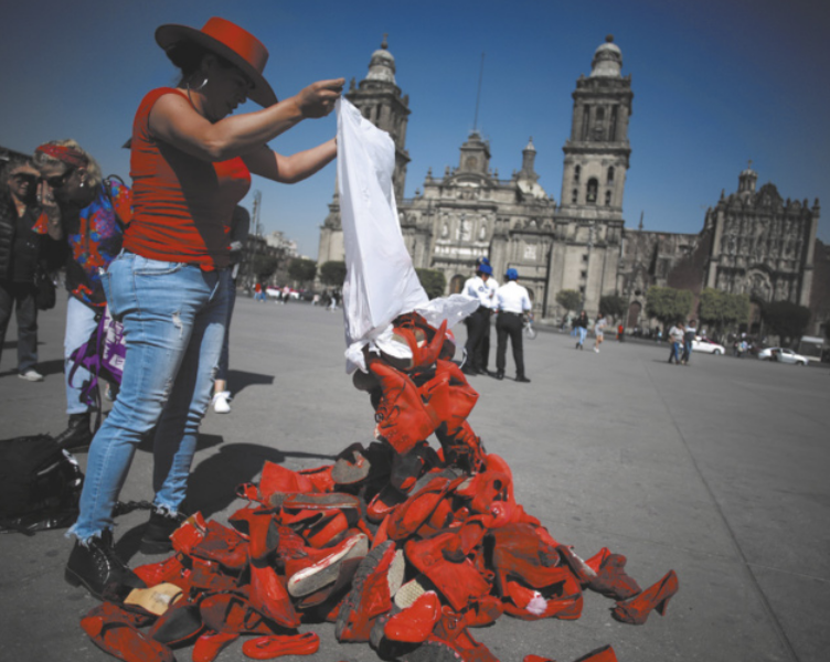 Fotografia de Elina Chauvet. Ela está em uma praça em frente a uma catedral, usando um chapéu e roupas vermelhas. Ela despeja uma pilha de sapatos vermelhos no chão.