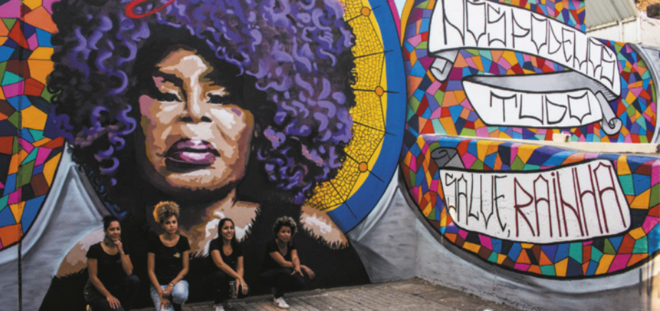Fotografia de quatro mulheres agachadas em frente a um muro com um grafite pintado, representando a cantora Elza Soares como uma mulher idosa com batom e cabelos cacheados de cor roxa. Ao lado do retrato de Elza Soares está escrita a mensagem: 'Nós podemos tudo. Salve, rainha'.
