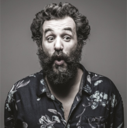 Fotografia de Alexandre Orion: ele é um homem com cabelos cacheados grisalhos e a barba longa, com a boca aberta, usando uma camisa de estampa florida.