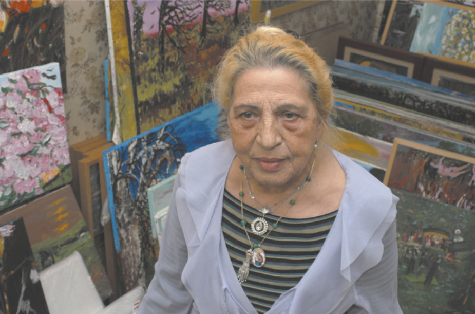 Fotografia de Ceija Stojka. Ela é uma mulher idosa com cabelos louros, usando um colar. Ela está ao lado de diversas pinturas apoiadas no chão e nas paredes de uma sala.