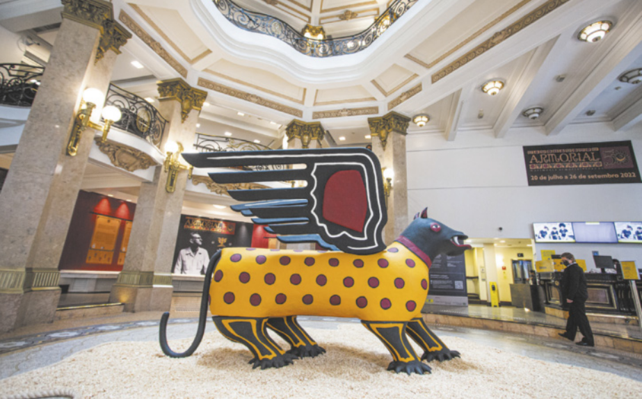 Fotografia de escultura representando um ser fantástico semelhante a uma onça-pintada com asas, com a superfície do corpo enfeitada com círculos vermelhos. A escultura está exposta no centro do salão de um edifício luxuoso com superfícies de mármore e ornamentos dourados.